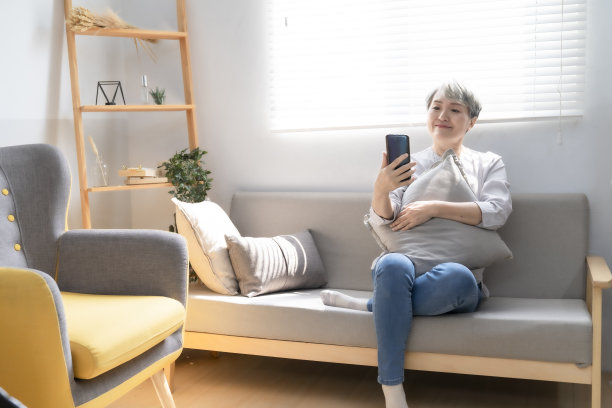 一位老妇人躺在家里客厅的沙发上，用智能手机和孙子进行视频会议。享受时光居家生活的高级家庭理念。人像照相机图片下载