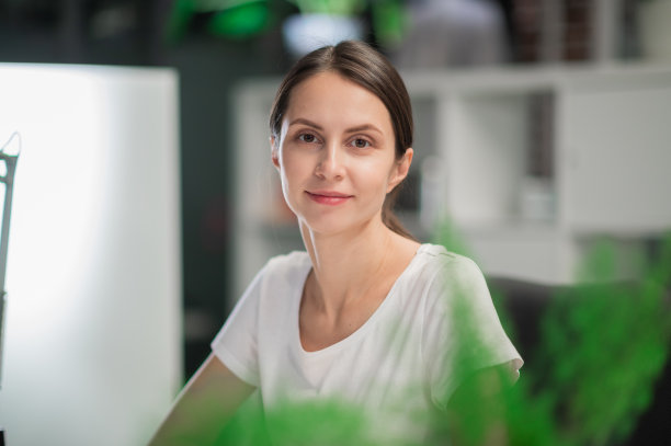 一个穿着白色t恤的年轻女子的肖像，看着镜头，坐在一张放着植物的桌子旁图片下载