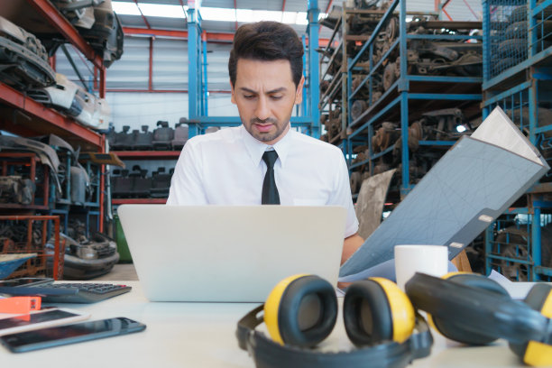 白种业务经理坐在工厂仓库里，拿着文件，用笔记本电脑认真工作图片下载