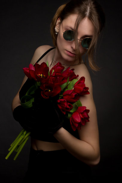 一个戴着墨镜的年轻女子的画室肖像，以黑色为背景，手里拿着一束红色郁金香图片下载