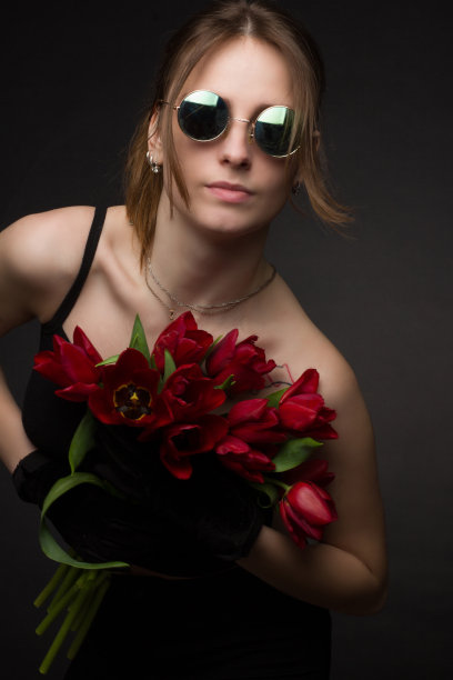 一个戴着墨镜的年轻女子的画室肖像，以黑色为背景，手里拿着一束红色郁金香图片下载