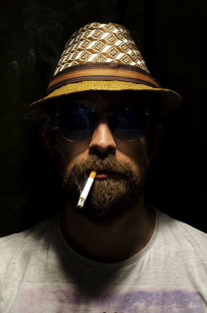 一个戴着太阳镜和帽子的大胡子男人的肖像。那人抽着烟。在黑色的背景上。图片下载