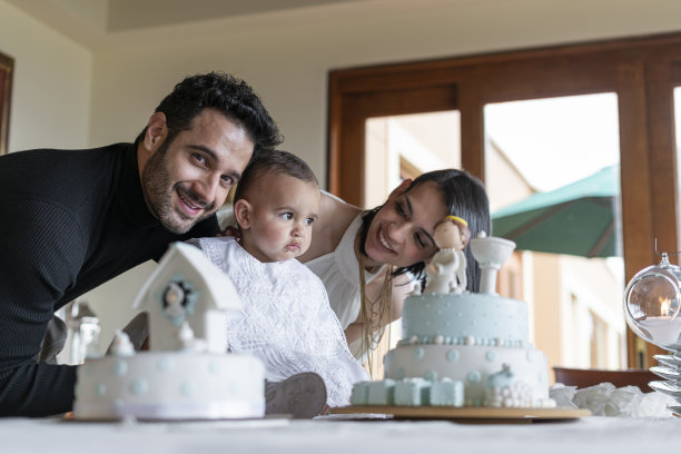 一对父母和他们的宝宝一起享受洗礼仪式，宝宝坐在桌子旁边，旁边是蛋糕，而骄傲的父亲在照片中看着相机图片下载