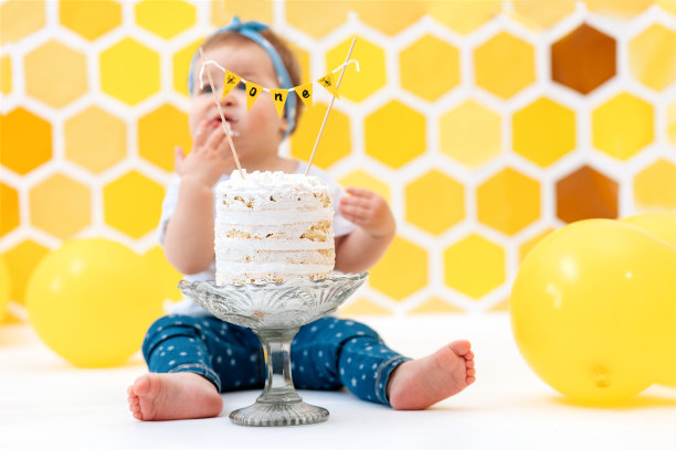 第一个生日。一个模糊的蹒跚学步的孩子坐在一块蛋糕旁边，用手吃一块。背景是黄色的蜂巢和气球的图案图片下载
