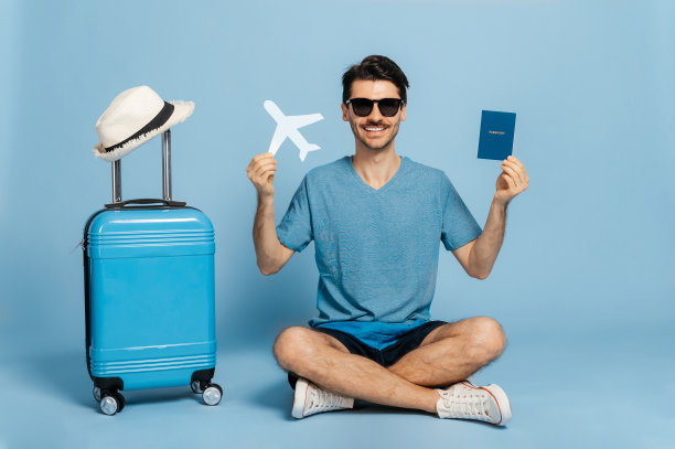 休假时间。快乐的白人男性游客戴着太阳镜和夏装坐在孤立的蓝色背景上，一手拿着护照，一手拿着飞机，蓝色行李箱站在旁边，微笑着图片下载