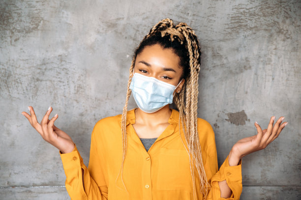 医疗保健。一个梳着脏辫的非洲裔美国女孩，穿着休闲衬衫，站在孤立的背景上，脸上戴着医用防护口罩，直视着镜头，用手打手势图片下载