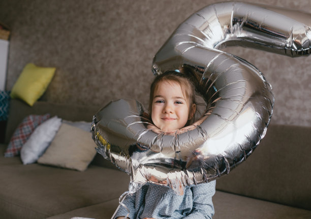 一个快乐的女孩在她的生日坐在沙发上，手里拿着一个巨大的充气闪亮的数字。5年。图片下载