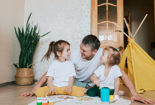 一位慈爱的年轻父亲和他的女儿们一起学习绘画，一个男人躺在地板上教女孩们画花，画家具。图片下载