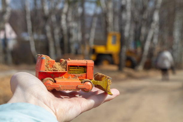 手上拿着一个推土机的玩具模型，背景是一辆黄色的真正的汽车图片下载