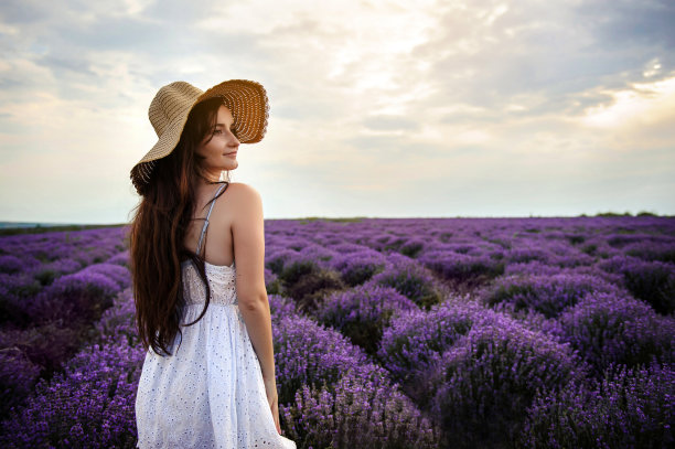 一个年轻的女人穿着白色的衣服和草帽走在薰衣草田的背影。图片下载