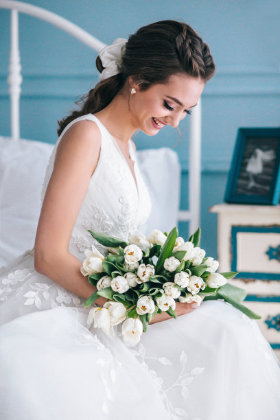 美丽的新娘带着婚礼花束坐在室内的床上，室内就像在家里一样。图片下载