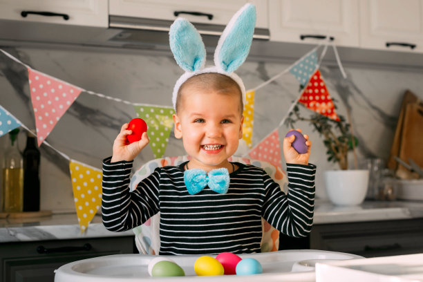 有趣的长着兔耳朵的金发男孩坐在厨房的桌子上玩彩色的复活节彩蛋。假期的概念。图片下载