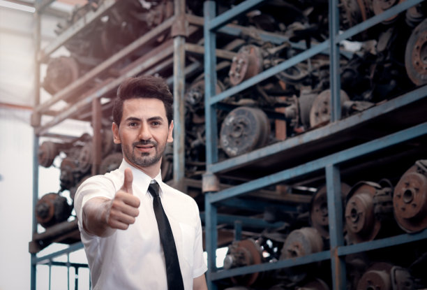 肖像微笑的商人卖家站在和给予拇指在汽车配件商店商店仓库与许多二手发动机零件模糊的背景。图片下载
