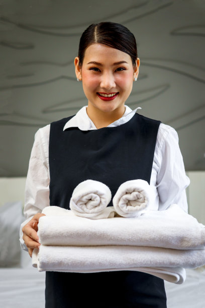 客房服务女佣打扫和整理床酒店房间的概念，年轻美丽的亚洲微笑女客房服务员的肖像抱着一叠新鲜的白色浴巾在酒店房间整理床。图片下载