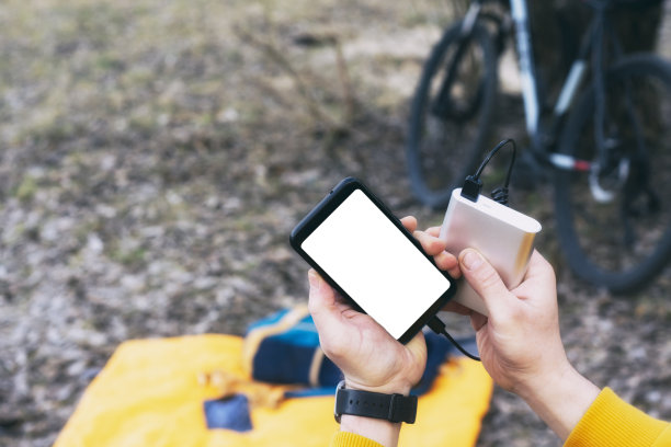 骑自行车的人手里拿着一个白色屏幕的智能手机模型，向银行收费。便携式充电器在自然界的背景上的自行车。图片下载