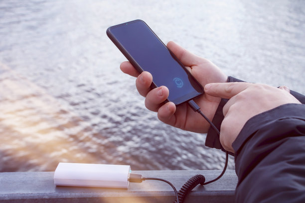 一名男子用充电棒给智能手机充电。在湖的背景下，手里的手机正在用便携式充电器充电。图片下载