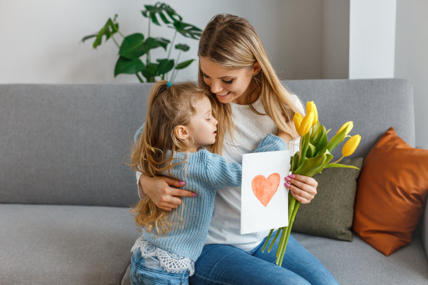 国际妇女节和母亲节。可爱的小女孩手里拿着一束黄色的鲜花和明信片，祝贺她微笑的妈妈。在家过一个快乐的家庭假期。卷发女孩抱着妈妈。图片下载