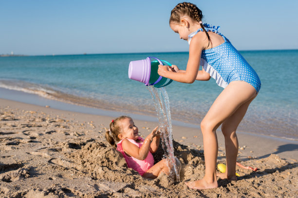 两个快乐的孩子在海滩上玩耍。女孩从桶里往姐姐身上倒水。在海边玩耍的孩子们。家庭度假的概念图片下载