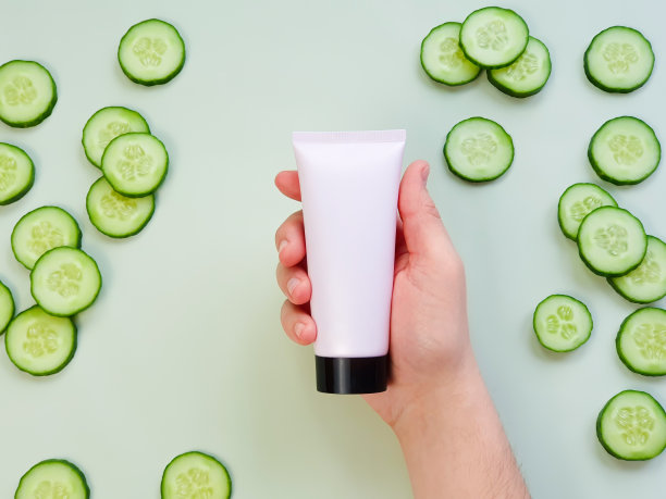 男性手拿塑料无商标的粉红色奶油管与黑色盖。瓶商标和标签和新鲜黄瓜切片在绿色背景。天然有机水疗化妆品。护肤品、广告图片下载