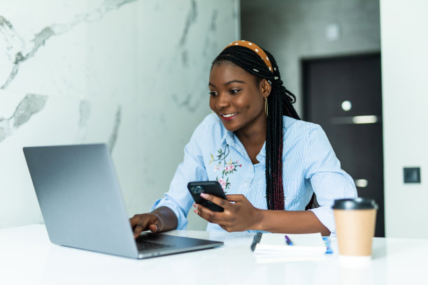 图片上的非洲女人微笑着卷着头发拿着智能手机在网上购物，付款，坐在厨房，使用笔记本电脑图片下载