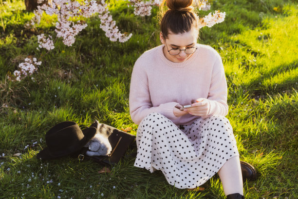 年轻的白人时髦女孩戴着眼镜，穿着浅色衣服，坐在公园里盛开的树附近的绿色草地上，使用她的智能手机。女人发短信，刷社交网络，聊天，看视频。图片下载