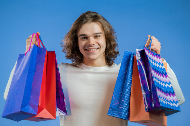 英俊的男人与长发持有购物纸袋在蓝色的工作室背景。那家伙买了打折的礼物。图片下载