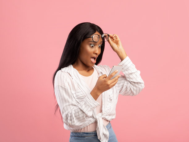 震惊的黑人女士看着智能手机难以置信，举起她的眼镜在粉红色的工作室背景图片下载
