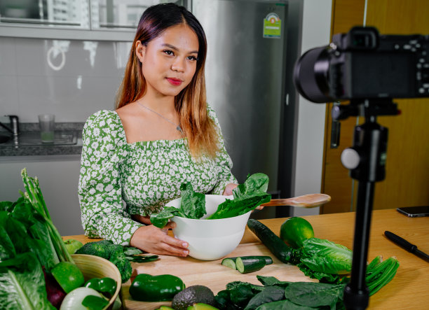 亚洲女性博主或内容创作者烹饪和录制摄像机。在厨房用摄像机拍摄的同时展示健康食品图片下载