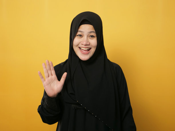 戴着头巾看着镜头微笑并挥手的穆斯林妇女，打招呼或告别手势，黄色背景图片下载