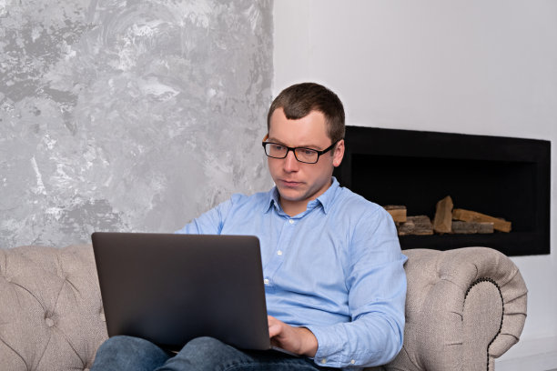 一位年轻的成功男士，穿着衬衫，戴着眼镜，坐在一间宽敞的办公室里，坐在沙发上，背靠壁炉，用笔记本电脑工作。成功与时间管理的理念。图片下载