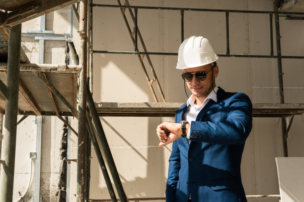 一个建筑师穿着正式的西装和安全帽看他的手表在一个建筑工地图片下载