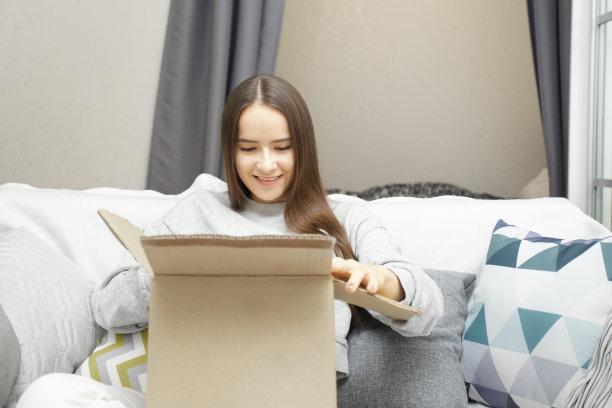 女孩坐在家里的沙发上，拿着一个纸箱打开包裹，开心地，微笑着图片下载
