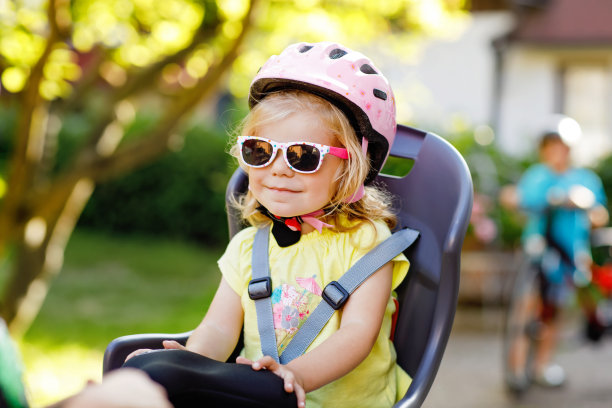 小女孩戴着安全帽坐在父母的自行车座位上的肖像。男孩骑自行车的背景。积极运动家庭休闲。家庭和周末活动旅行。图片下载