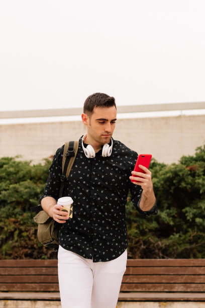 一个英俊的年轻人用他的智能手机在街上喝咖啡的垂直照片。生活方式的概念。图片下载