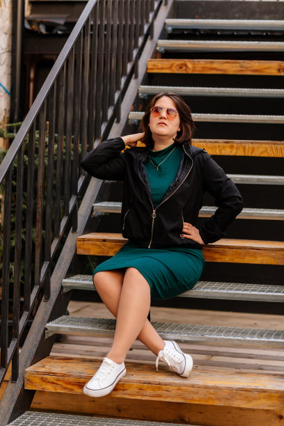 时髦的年轻漂亮的潮女人坐在铁房子的楼梯上，快乐美丽的女孩在休闲衣服绿色连衣裙，黑色夹克，白色运动鞋，城市背景，街头照片，身体正能量图片下载
