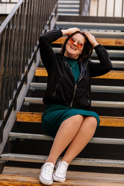 时髦的年轻漂亮的潮女人坐在铁房子的楼梯上，快乐美丽的女孩在休闲衣服绿色连衣裙，黑色夹克，白色运动鞋，城市背景，街头照片，身体正能量图片下载