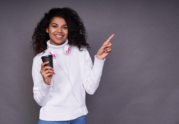 年轻漂亮的黑皮肤女孩拿着咖啡向一边伸出手指。图片下载