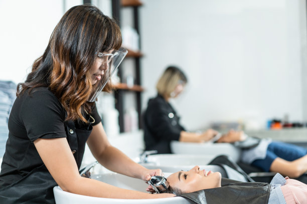 亚洲女性美发师，在新冠肺炎流行期间戴面罩，为顾客按摩头发和用洗发水洗头。在现代沙龙理发店服务时，练习社交距离图片下载