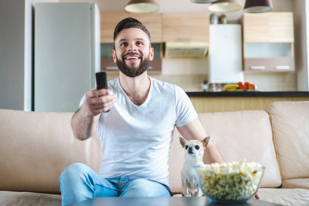 一幅年轻英俊的大胡子男子周末在家坐在沙发上，和他的小朋友吉娃娃狗一起吃爆米花看电视的照片图片下载