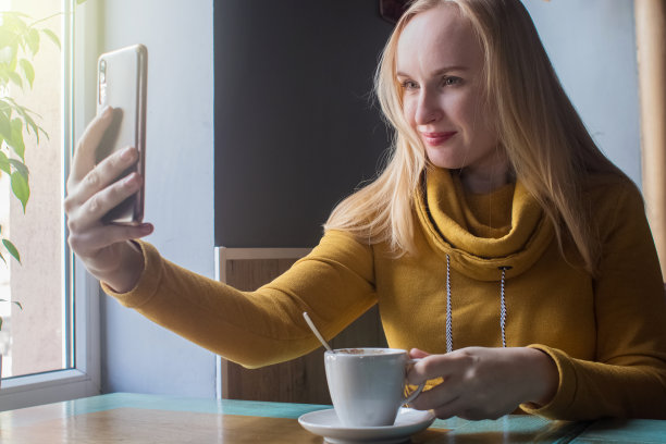 博主女孩坐在咖啡馆里用智能手机拍一杯卡布奇诺泡沫。在社交网络、视频博客和照片博客中工作。互联网内容创作和食物摄影的概念。图片下载
