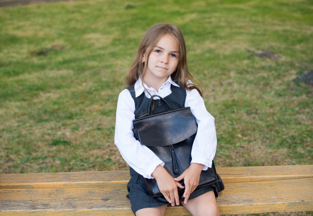 可爱的小孩子穿着正式的校服，神情严肃地拿着书包坐在公园长椅上，准备上学图片下载