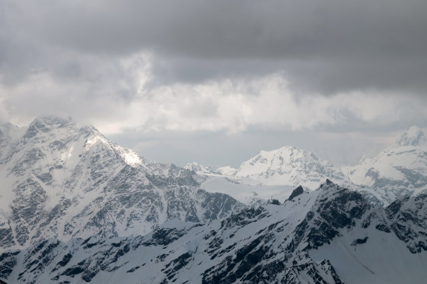 层层叠叠的蓬松的云高高地在积雪覆盖的山峰上，光线穿过它们。山tele景观。非对比性背景的旅行和横幅图片下载