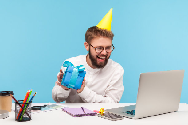 一名面带微笑的白领戴着圆锥形的帽子打视频电话，在笔记本电脑的网络摄像头上展示蓝色的礼盒，祝贺网上彩票的中奖者图片下载