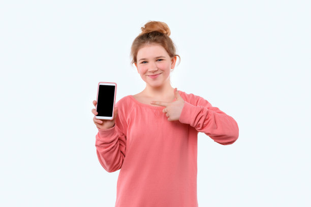 一个穿着粉色休闲运动衫的可爱小女孩拿着智能手机，用左手食指指着它图片下载