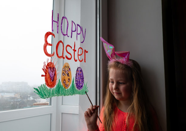 可爱的女孩戴着兔子耳朵在窗户上画复活节彩蛋绿草。孩子准备画节日的家饰图片下载