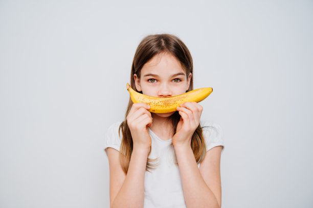 一个可爱的十几岁的女孩拿着香蕉对着她的脸，对着一堵白色的墙微笑。图片下载