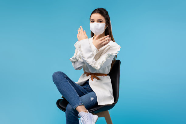 一名身穿白色衬衫、戴着医用口罩的年轻女孩坐在办公椅上，蓝色背景显示的是“停止”，即病毒传播的终结。工作和流行。图片下载
