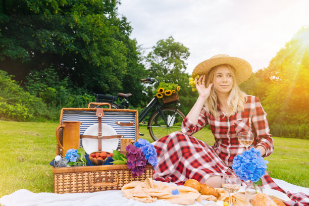 女孩穿着红色格子裙和帽子坐在白色针织野餐毯上看书和喝葡萄酒。在阳光明媚的夏日野餐，有面包，水果，绣球花。图片下载