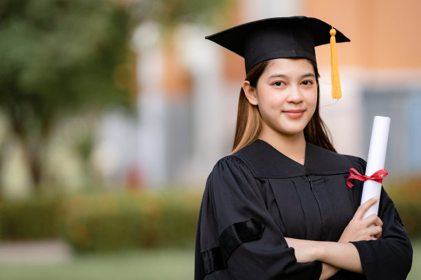 一位年轻快乐的亚洲女子大学毕业生穿着毕业礼服和学位帽持有学位证书庆祝教育成就在大学校园。教育库存图片图片下载