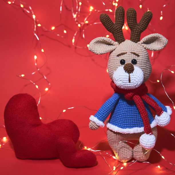 可爱的背景为情人节。一只迷人的手工小鹿在蓝色毛衣和红色针织围巾在红色的背景与灯花环和两颗心。图片下载